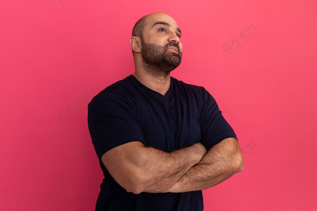 立场一个留着大胡子的穿海军t恤的男人 双臂交叉着站在粉红色的墙上 神情严肃地沉思着严肃表情男人