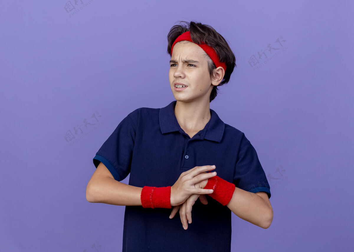 牙科皱眉的年轻帅气的运动男孩戴着头带和护腕 戴着牙套 一边看一边把手放在另一只手上 在紫色的墙上练习 有复制空间戴运动头带