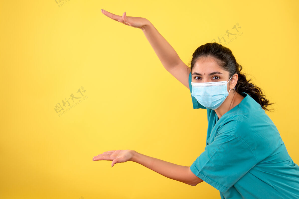 快乐戴着医用面罩的年轻女医生的正面图 显示了手放在黄色墙上的尺寸肖像年轻女医生秀