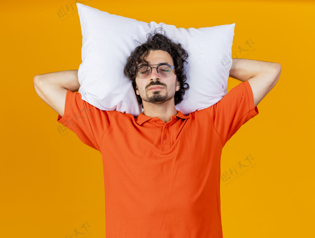 头昏昏欲睡的年轻白种人 戴着眼镜 头下抱着枕头 在橙色的墙上假装闭着眼睛睡觉穿睡觉生病