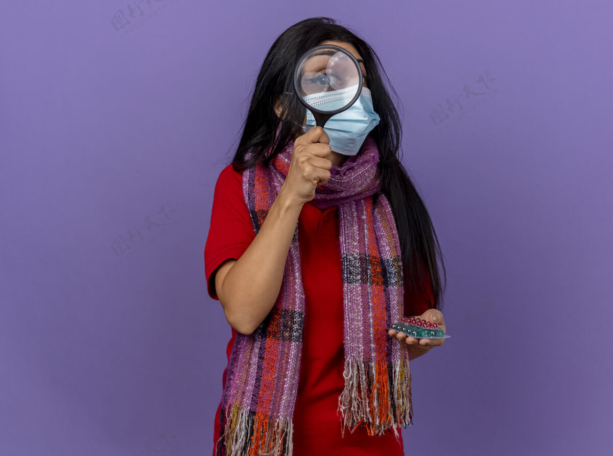 包装年轻的白种人患病的女孩戴着口罩和围巾 手里拿着一包包胶囊 通过放大镜隔离在紫色的墙上 留着复印空间玻璃穿过围巾