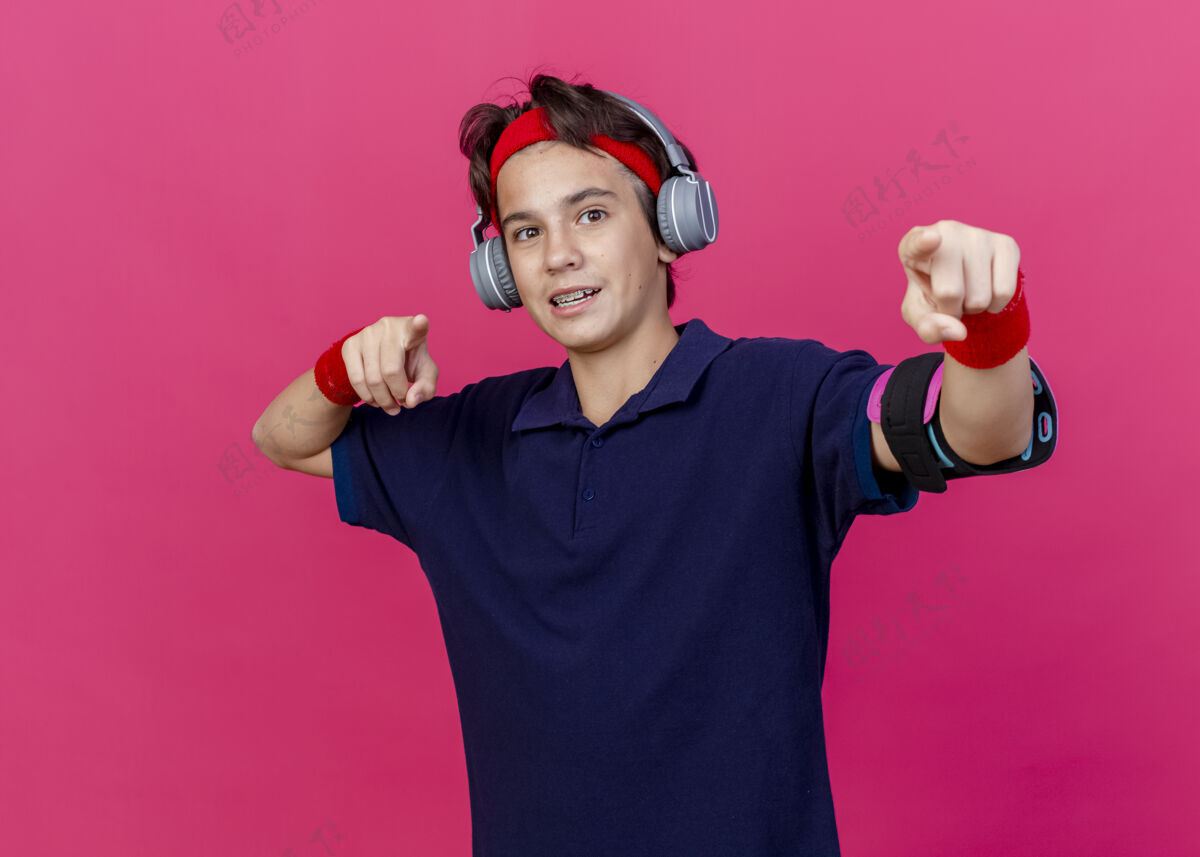 背带令人印象深刻的年轻英俊的运动男孩戴着头带和腕带和耳机电话袖带与牙套看 并指出在深红色的墙上孤立袖带印象指尖