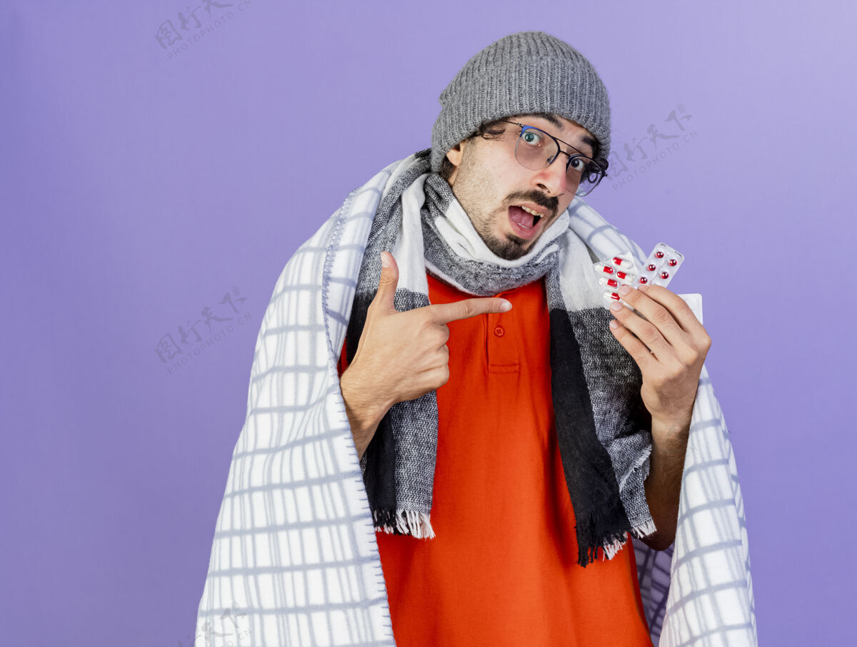 男人印象深刻的年轻白种人 戴着眼镜 戴着冬天的帽子 围着方格布围巾 拿着并指着紫色墙上孤立的一包包胶囊 还有复印空间帽子包穿