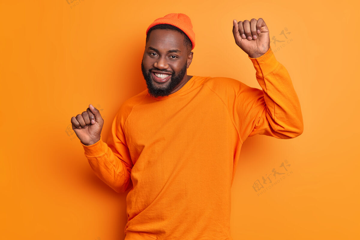 帽子快乐无忧无虑的家伙在橘色的墙上跳舞举手积极微笑高兴地戴上时髦的帽子和套头衫庆祝成功站立表情帅气