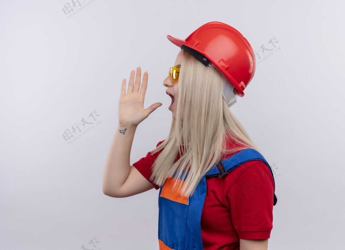 安全年轻的金发碧眼的建筑工女孩 穿着制服 戴着安全眼镜 站在隔离的白色墙壁上 对着有复印空间的人大声喊叫 手靠近嘴巴喊叫建设者手