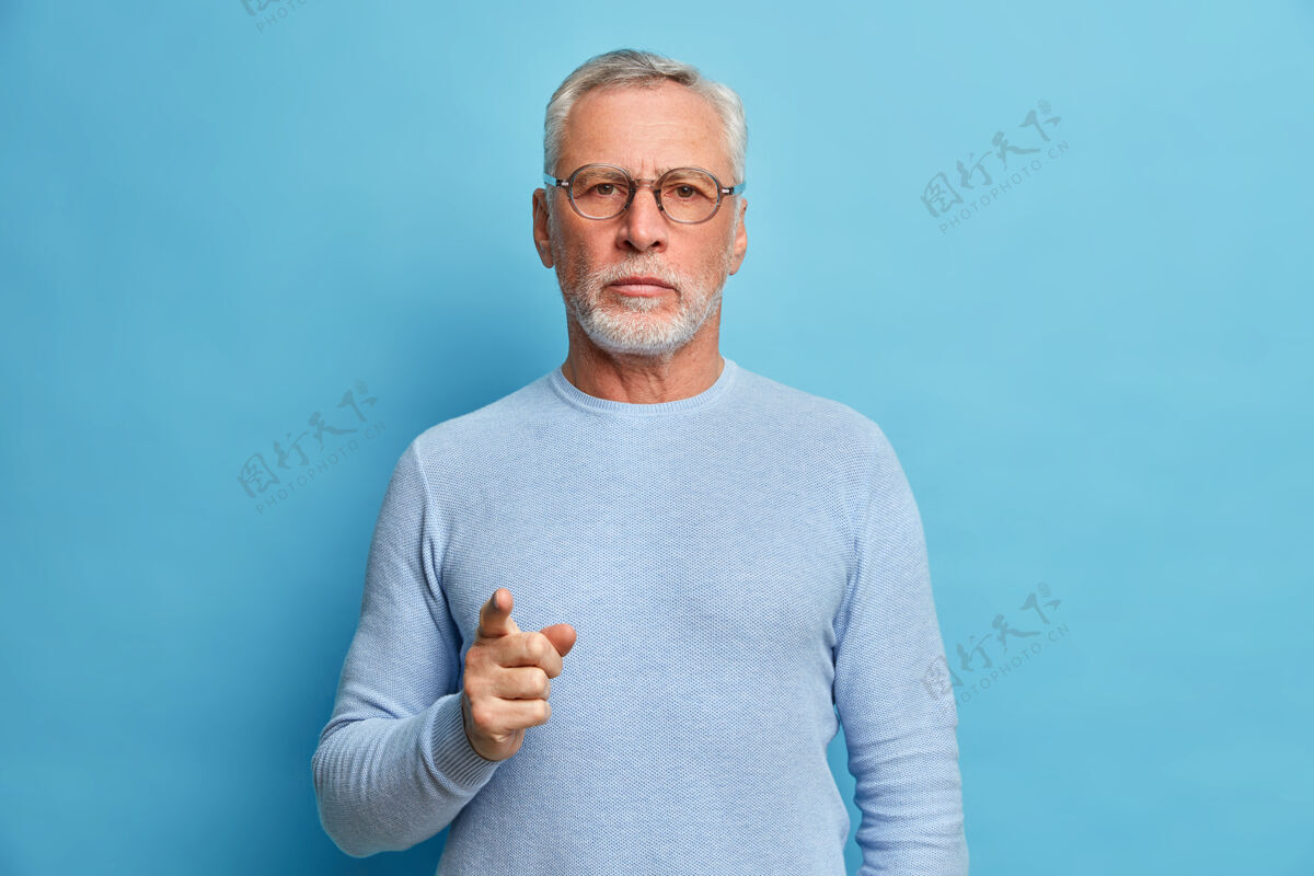 年龄自信成熟的胡须男人指在前面 选择你认真地看镜头选择一个人在他的团队穿休闲套头衫隔离蓝墙眼镜脸皱纹