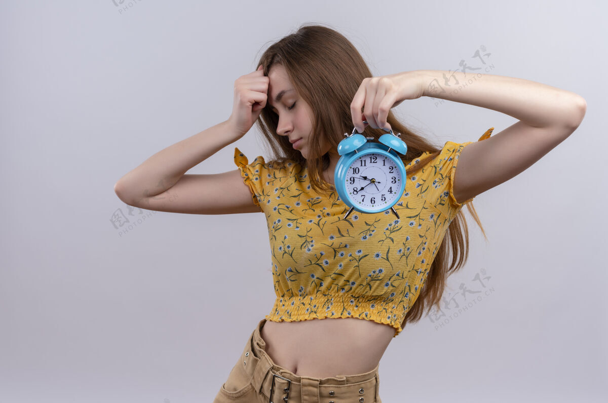 女孩疲惫的年轻女孩拿着闹钟 把手放在孤立的白墙上的额头上手累了年轻