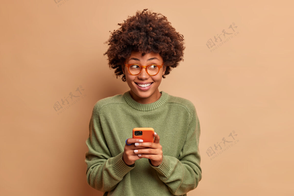 沟通漂亮女人的画像用手机收到愉快的信息笑容满面 一边若有所思地看一边在社交网络隔离的米色墙上阅读她的帖子下的反馈眼镜民族快乐