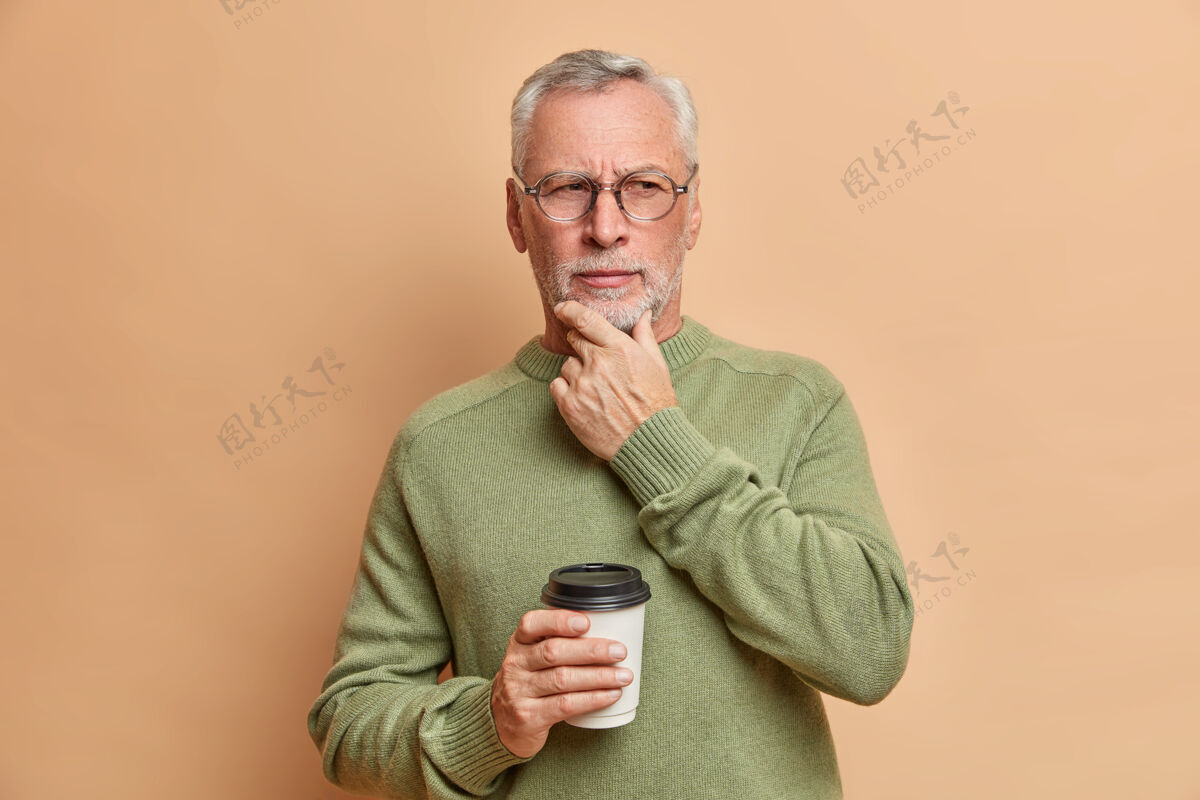 男非常严肃成熟的欧洲男人抱着下巴沉思地看着一边喝咖啡去考虑重要的事情穿休闲毛衣和眼镜隔着棕色的墙高级成熟眼镜