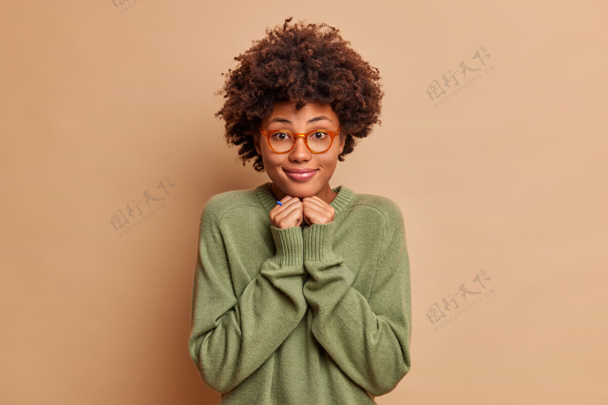 个人快乐开朗的女学生留着非洲式头发手放在下巴下穿着休闲套头衫戴着眼镜脸上露出迷人的笑容隔着米色的墙壁自然穿女性
