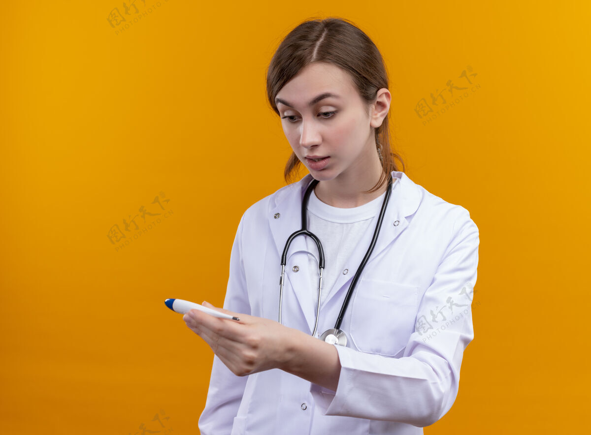 穿年轻的女医生穿着医用长袍 手持听诊器 拿着体温计 惊讶地看着隔离的橙色墙壁上有复印空间听诊器女惊喜