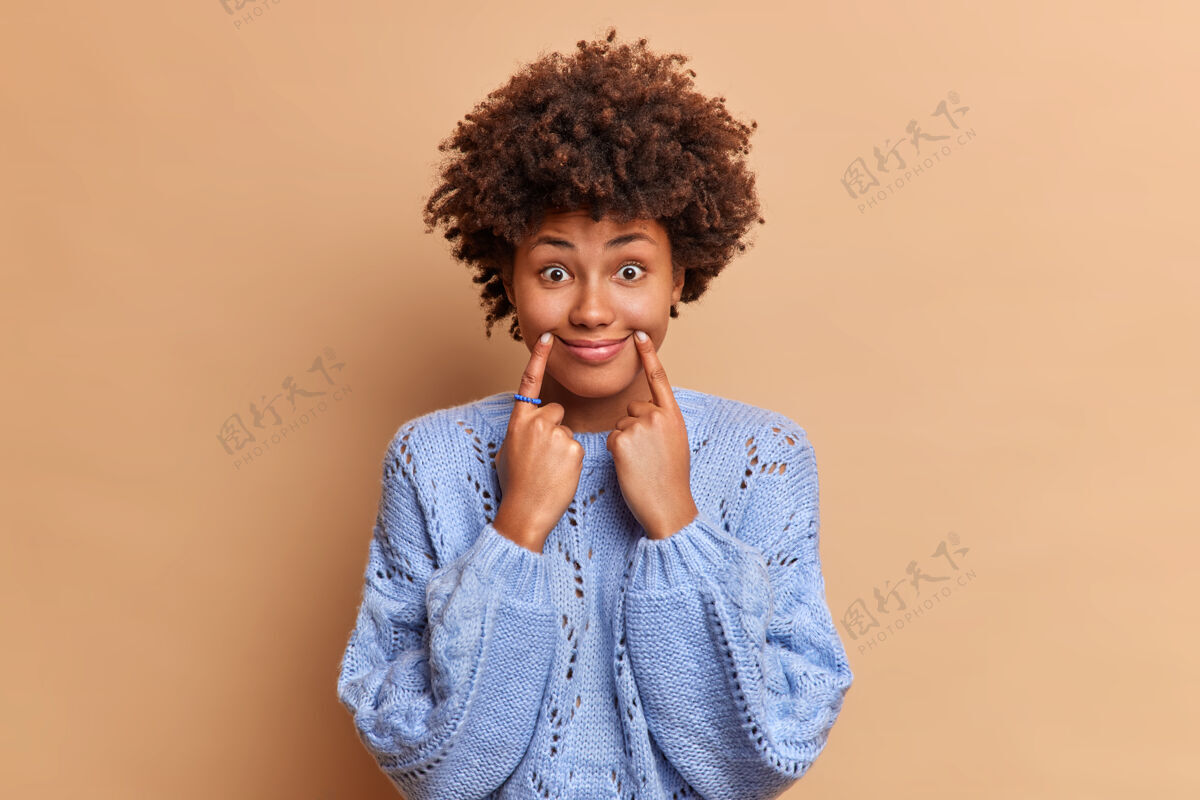 发型有趣的女人用手指制造微笑强迫自己有积极的表情穿着休闲毛衣靠着棕色的墙壁摆姿势民族心烦触摸