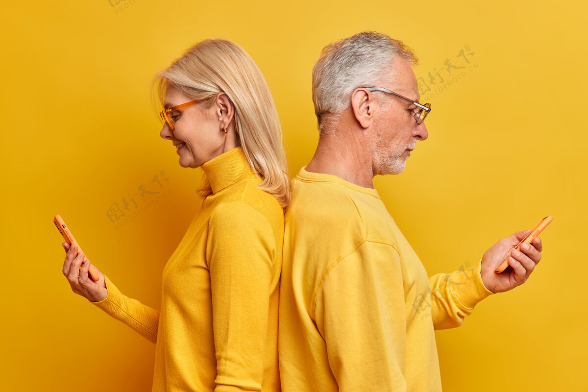 现代老妇人和老男人背靠背站在一起使用现代智能手机聊天上网冲浪享受家庭时光沉迷于技术隔着黄色的工作室墙请电话科技