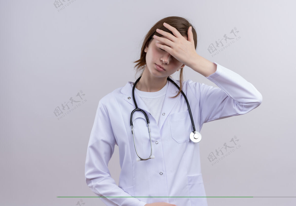 长袍压力大的年轻女医生穿着医用长袍和听诊器把手放在额头上 在隔离的白色墙壁上留有复印空间听诊器年轻人手