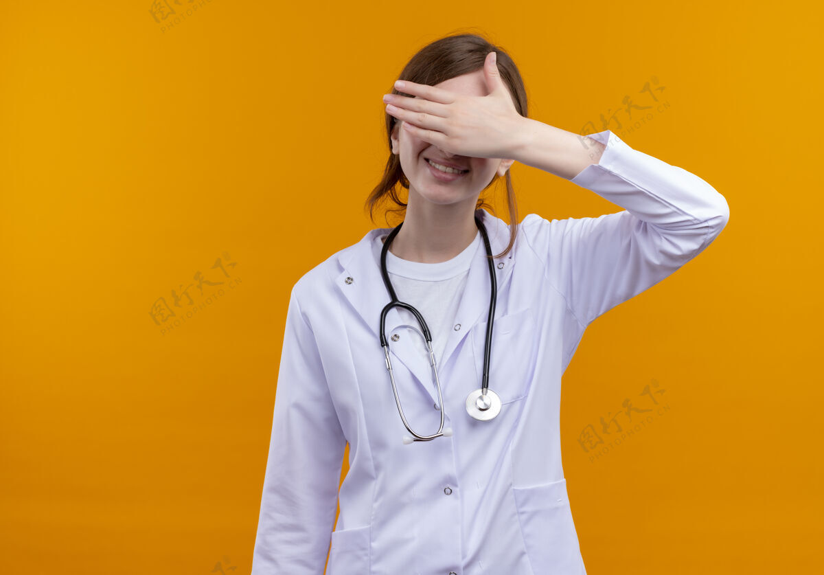 穿微笑的年轻女医生穿着医用长袍和听诊器闭上眼睛 手放在隔离的橙色墙壁上 留有复印空间年轻手医学