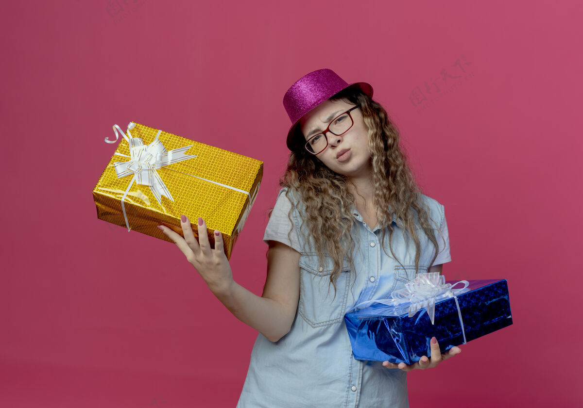 戴着戴着眼镜 戴着粉红色帽子 手里拿着礼品盒的年轻女孩被隔离在粉红色的墙上拿着女孩眼镜