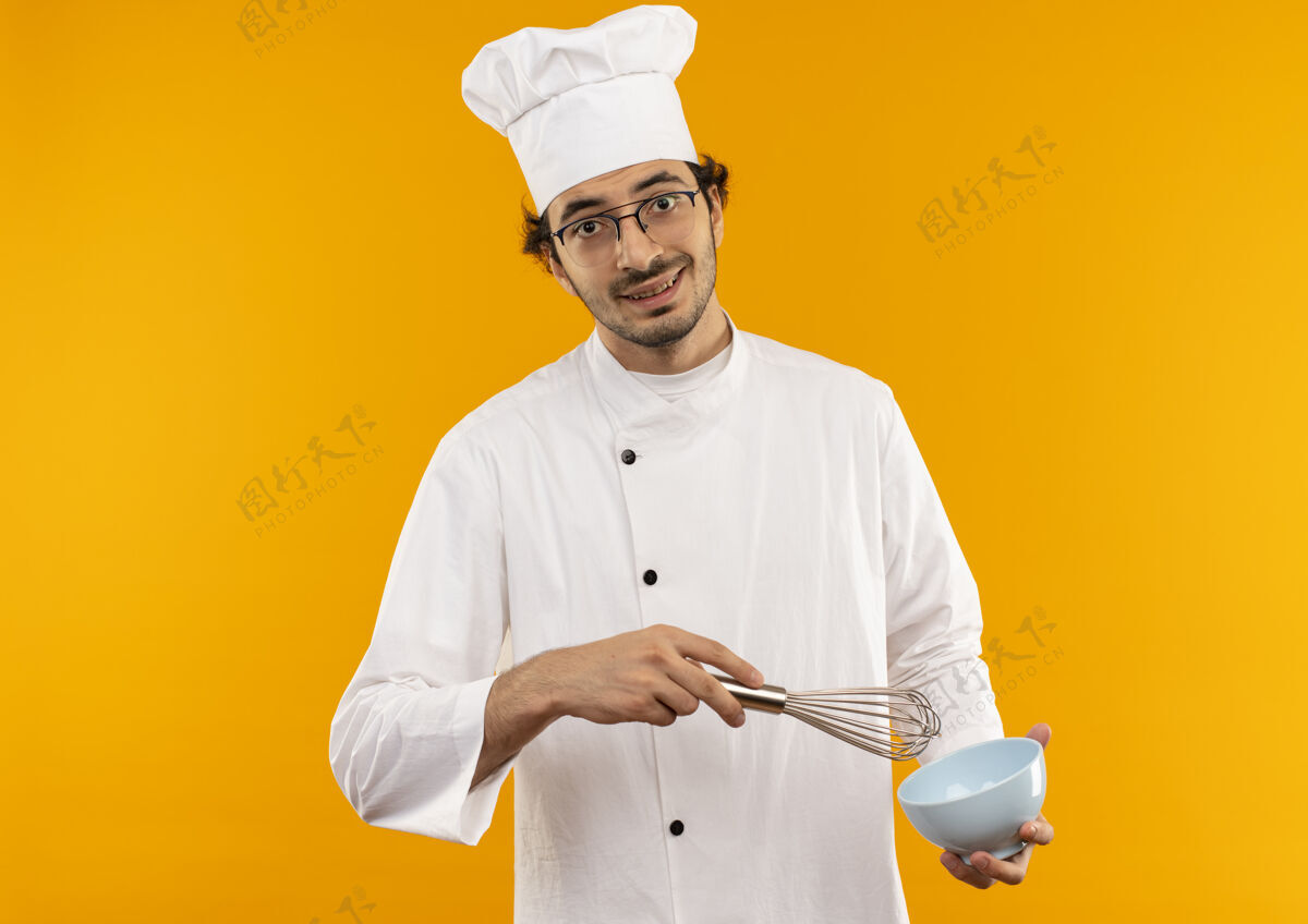 男高兴的年轻男厨师穿着厨师制服 戴着眼镜 拿着碗和威士忌隔离在黄色的墙上厨师眼镜烹饪