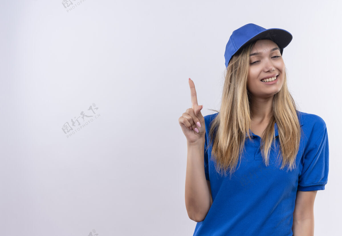 女孩带着微笑闭上眼睛的年轻送货员身穿蓝色制服 戴着帽子 指着隔离在白色墙上的复制空间年轻帽子微笑