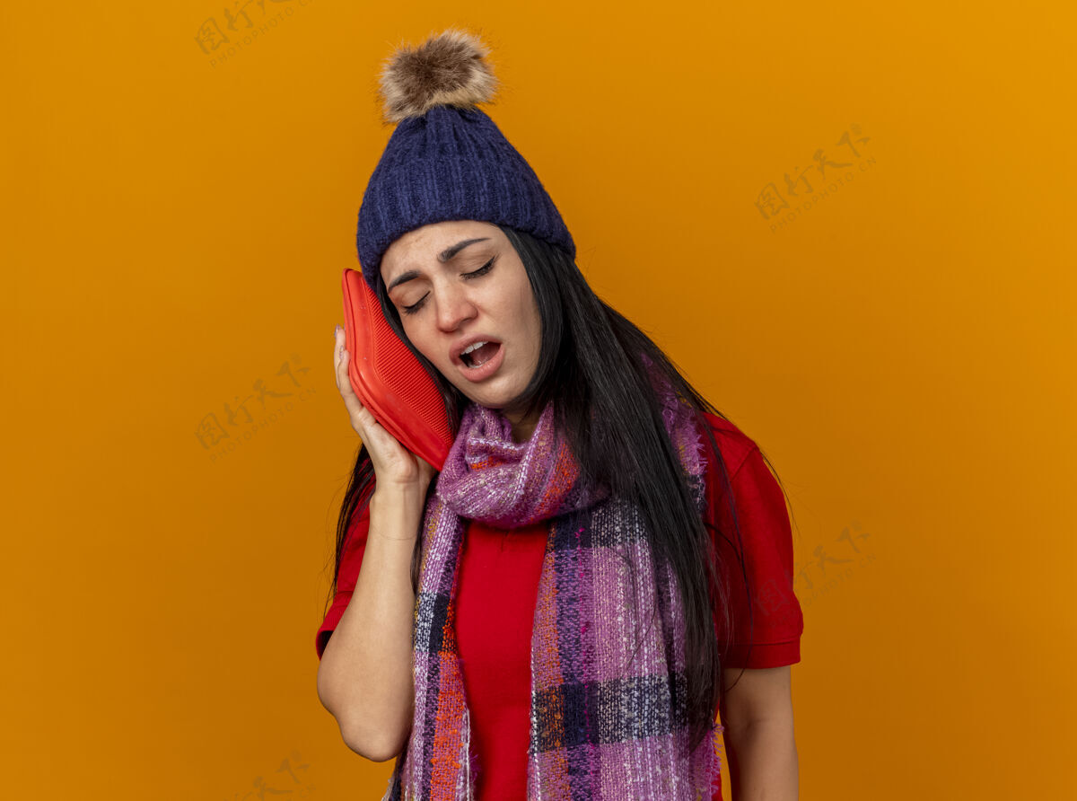年轻弱小的年轻白种人患病的女孩戴着冬天的帽子 戴着围巾 用热水袋摸着脸 闭着眼睛 隔离在橙色的墙上 留着复印空间疾病帽子橙色