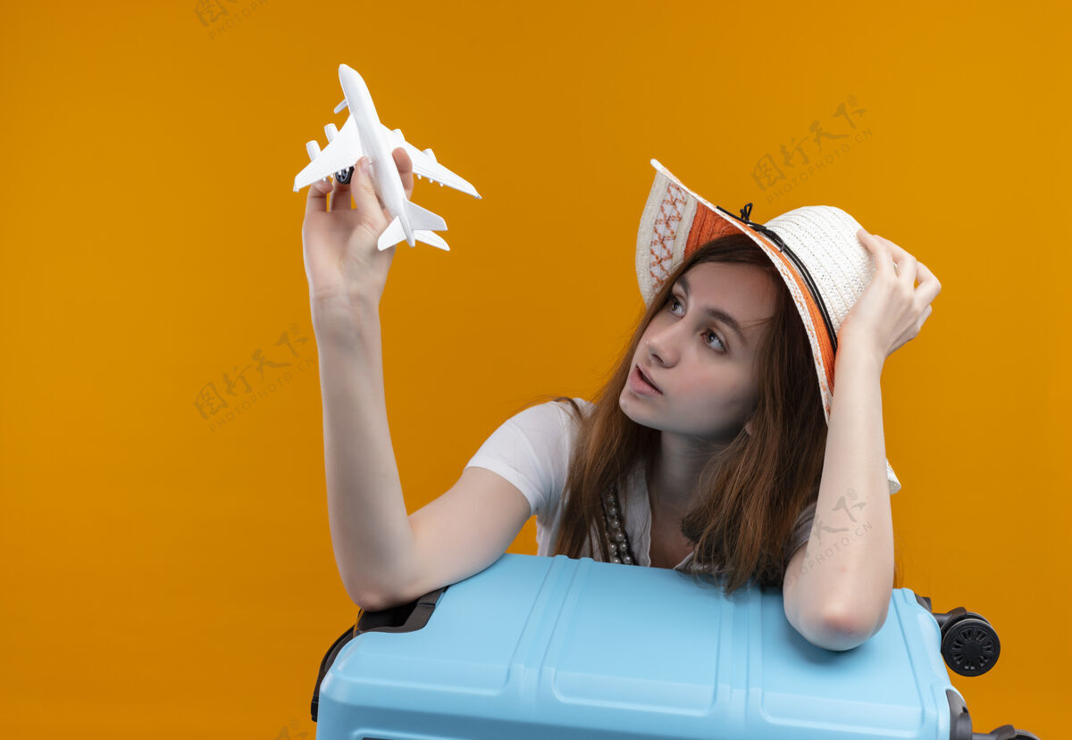 年轻人年轻的旅行女孩戴着帽子拿着模型飞机 看着它 把胳膊放在手提箱上 手放在帽子上 放在隔离的橙色墙壁上 留着复制空间戴着飞机女人