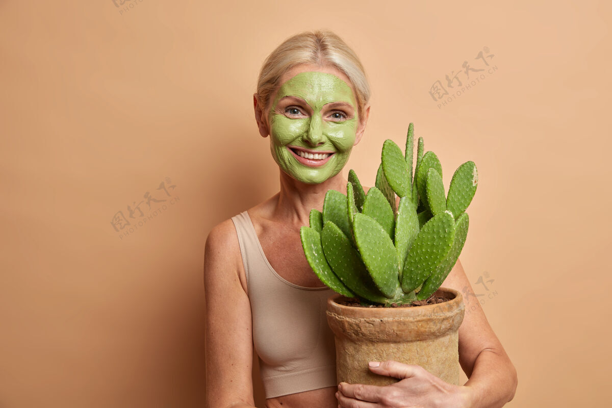 表情积极可爱的中年妇女关心皮肤涂绿色滋养面膜在脸上拥抱仙人掌微笑轻轻隔离在棕色墙壁锅人正面人