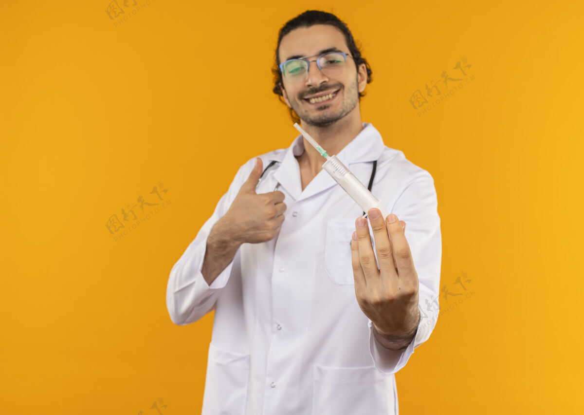 眼镜面带微笑的年轻医生 戴着医用眼镜 穿着医用长袍 手持听诊器 拿着注射器 他的大拇指竖立在隔离的黄色墙壁上 有复印空间年轻长袍拿着