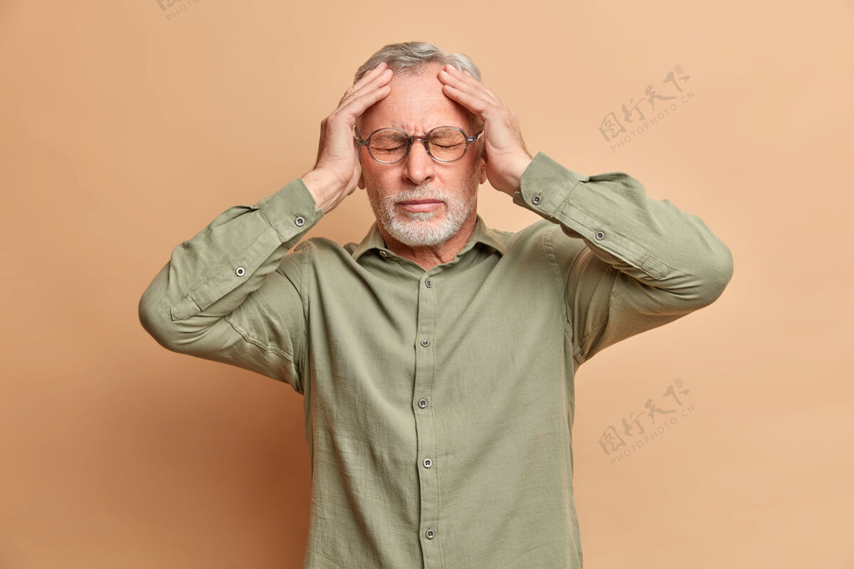人不高兴的白发男子头痛手放在头上透露疼痛需要止痛药有偏头痛喧闹派对后穿正式衬衫隔离在棕色墙壁上表达祖父老