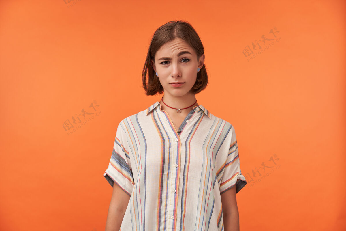 表情一个年轻的孤立无援的女人站在橙色的墙上看着你 扬起眉毛 穿着条纹衬衫当你听到一些奇怪的事情时成人女士悲伤