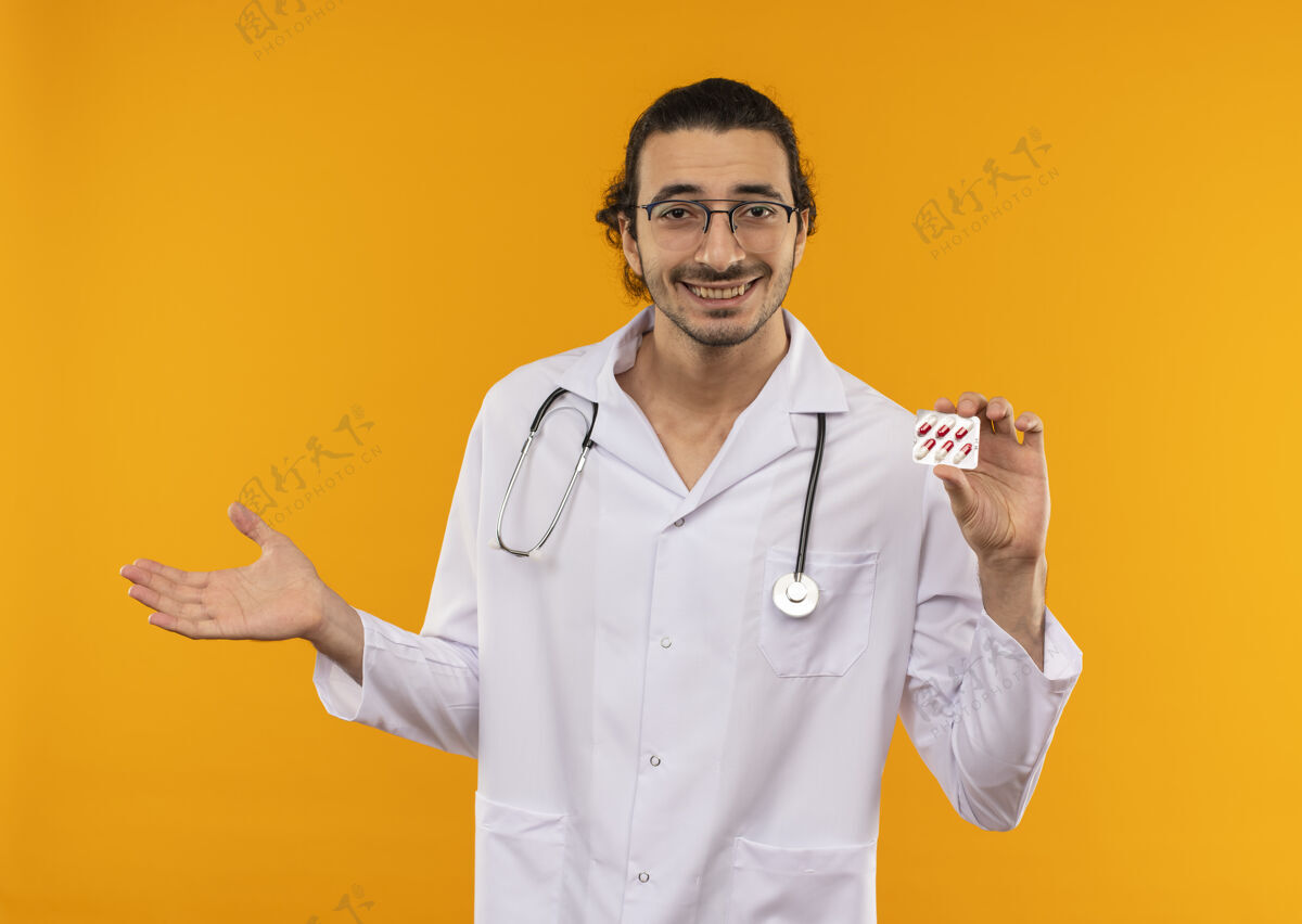 年轻面带微笑的年轻医生戴着医用眼镜 穿着医用长袍 手持听诊器 手持药片 手放在隔离的黄色墙壁上 留有复印空间长袍眼镜手