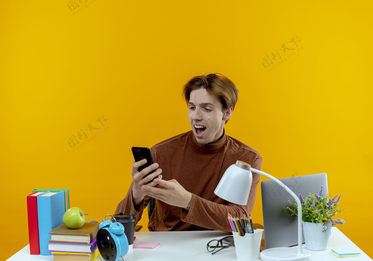 工具惊讶的年轻学生男孩坐在书桌旁 手里拿着学习工具 看着隔离在黄色墙上的电话抱着年轻人坐着