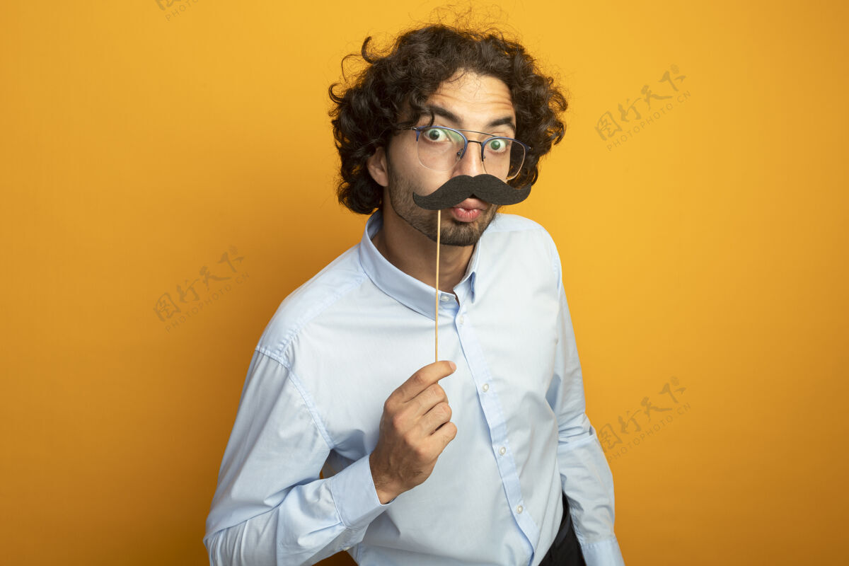 保持有趣的年轻英俊的白种人戴着眼镜留着假胡子贴在嘴唇上方看着摄像机做亲吻动作在橙色背景上用复制空间隔离胡子棒相机