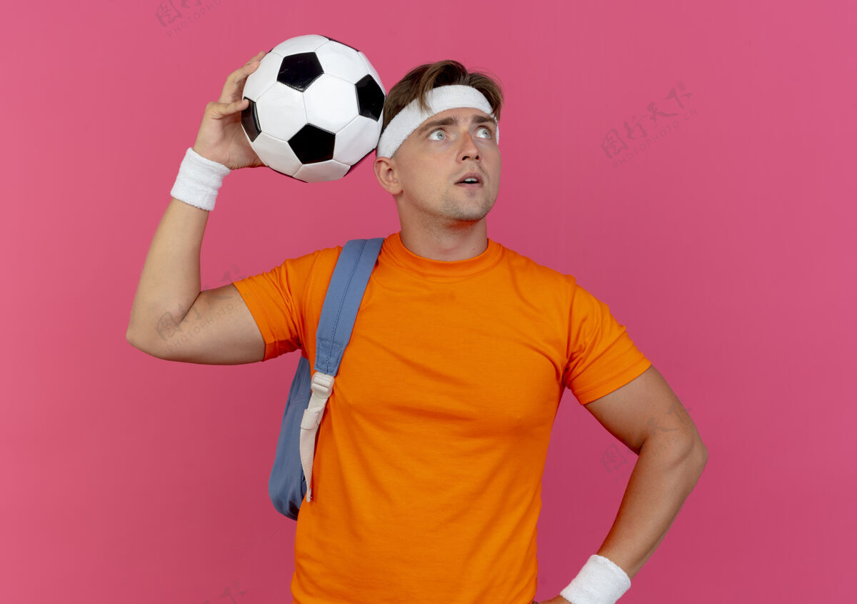 手令人印象深刻的年轻英俊的运动型男子戴着头带和腕带和背包拿着足球和触摸它的头上看与腰部隔离的手在粉红色运动向上背部
