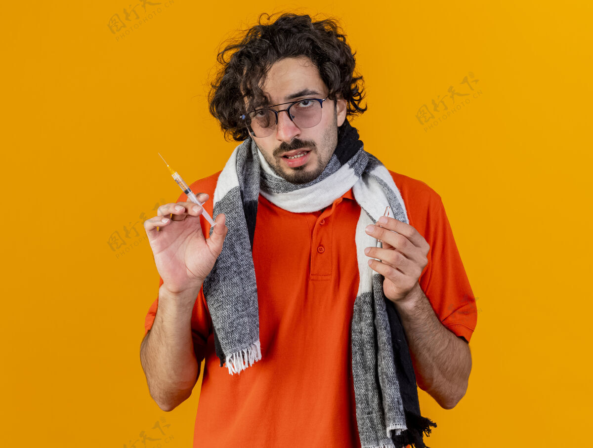相机年轻的白人病人戴着眼镜 戴着围巾 拿着注射器和安瓿 看着隔离在橙色背景下的摄像机持有疾病注射器