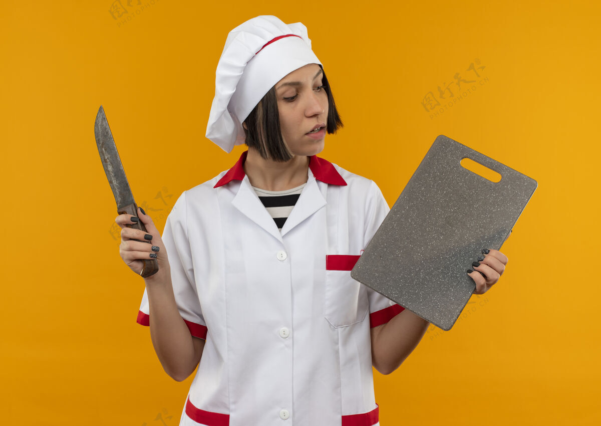年轻印象深刻的年轻女厨师穿着厨师制服拿着刀和砧板 看着板上孤立的橙色烹饪厨师橙