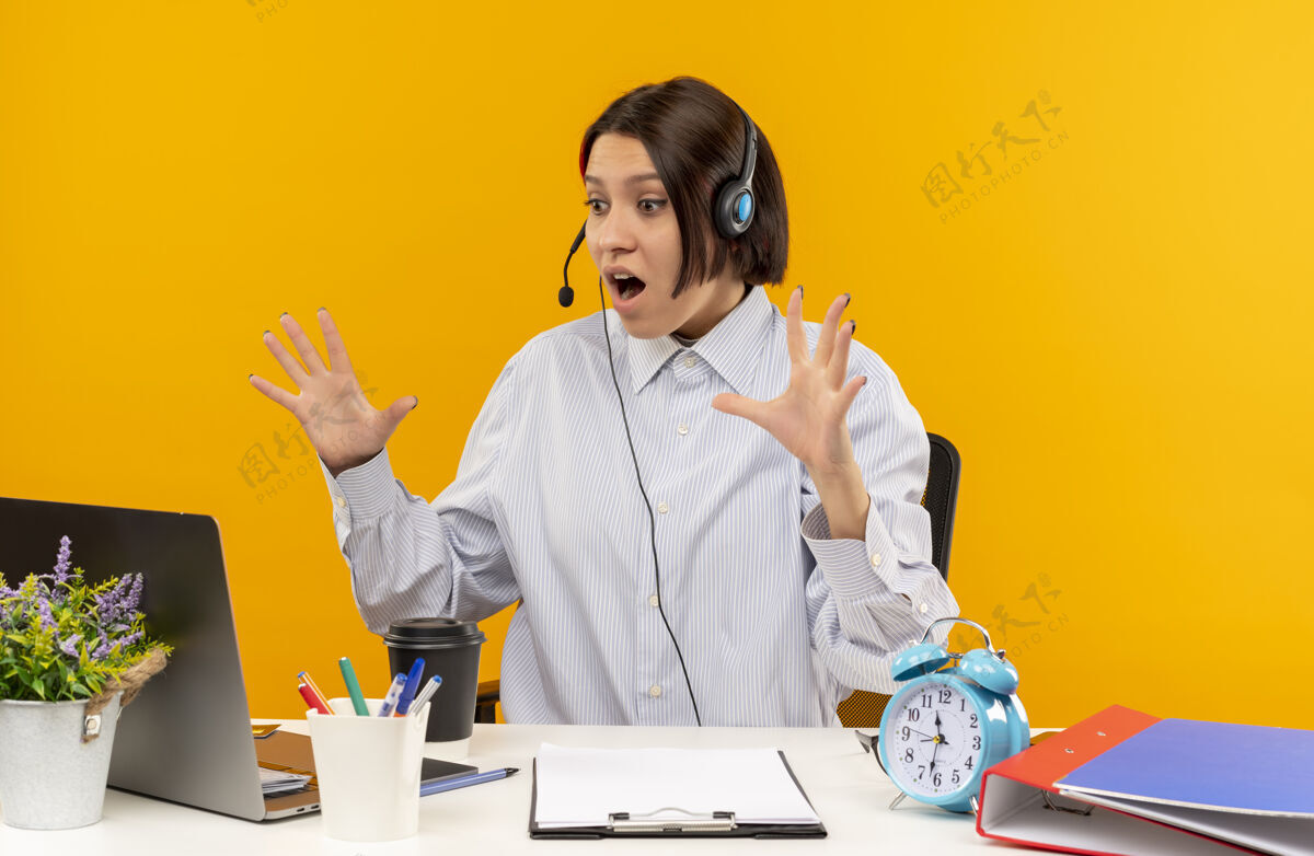 中心令人印象深刻的年轻呼叫中心女孩戴着耳机坐在办公桌旁看着笔记本电脑举起的手孤立在橙色年轻橙色印象