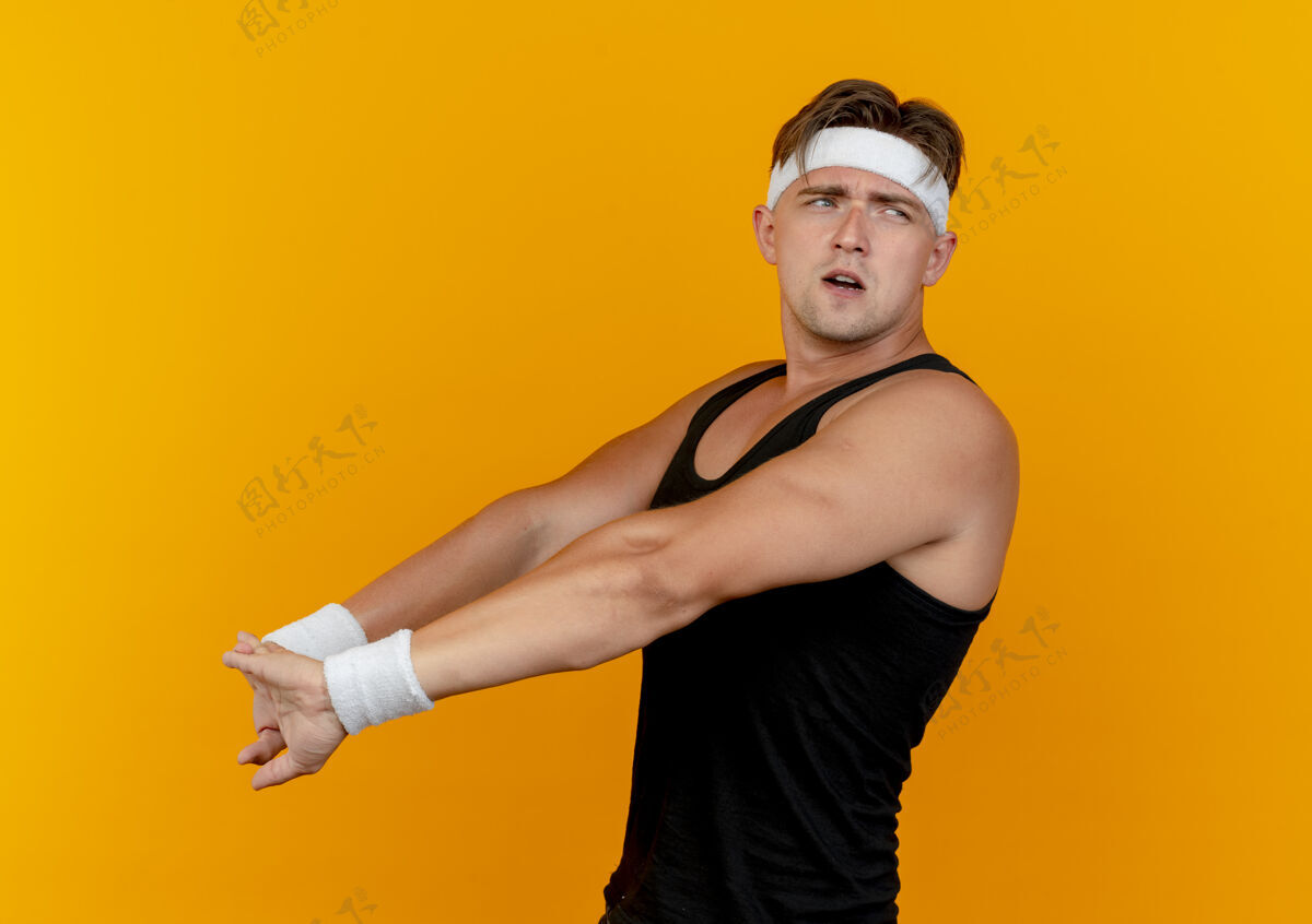 迷茫迷茫的年轻英俊的运动型男子戴着头带和腕带站在侧视图看边和伸展的手孤立在橙色站立运动腕带