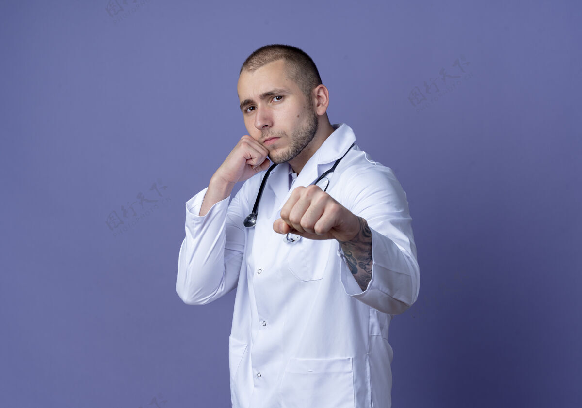 自信自信的年轻男医生穿着医用长袍和听诊器做拳击手势 在紫色的空间里与世隔绝手势年轻长袍