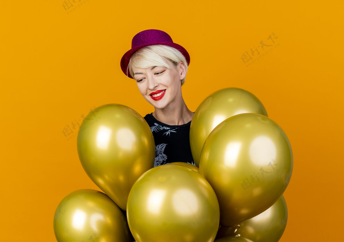 气球微笑着的金发派对女郎戴着派对帽站在气球后面看着橙色背景下孤立的气球微笑站着女孩