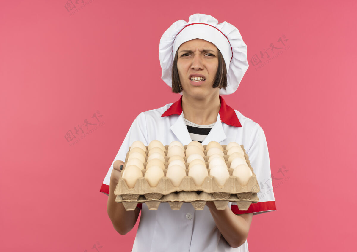 持有恼火的年轻女厨师穿着厨师制服 手里拿着一盒鸡蛋 粉红色的盒子上有复制空间年轻女纸箱