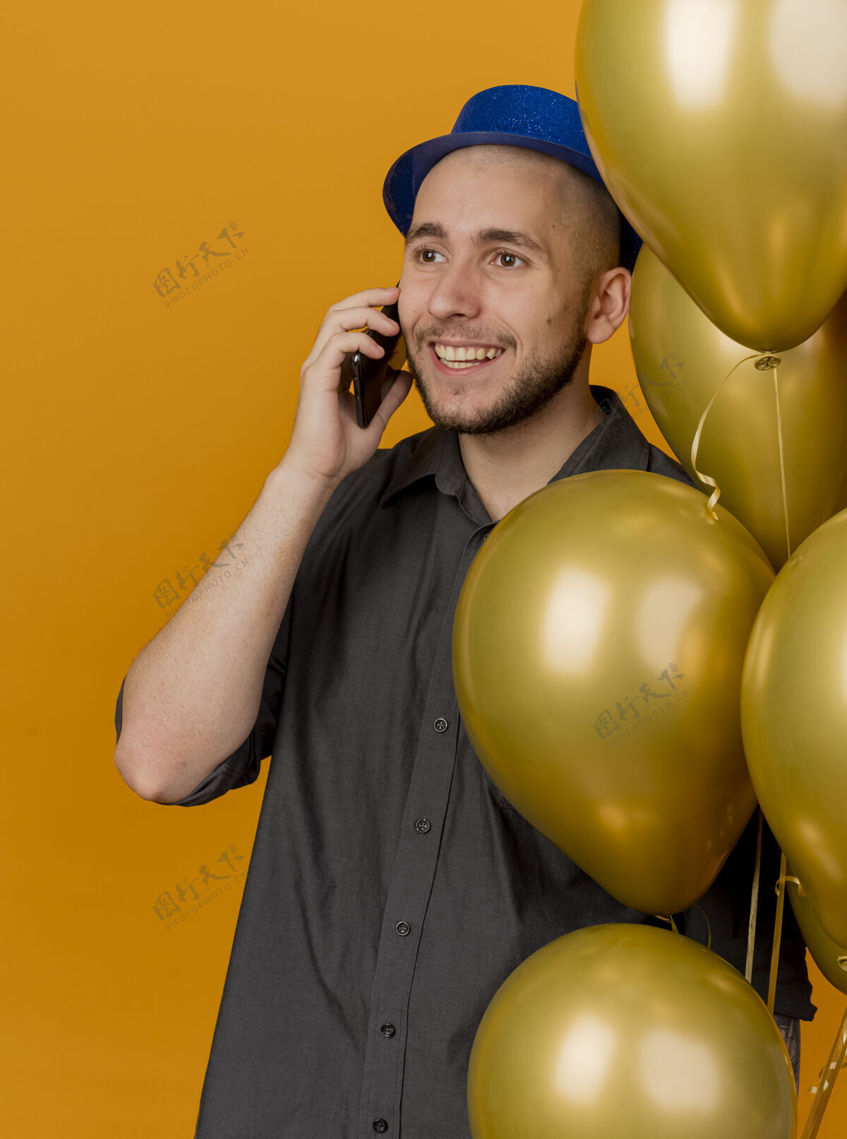 抱着面带微笑的年轻英俊的斯拉夫党人戴着党的帽子拿着气球在电话里说话看起来直挺挺孤立的橙色背景帽子小伙子电话
