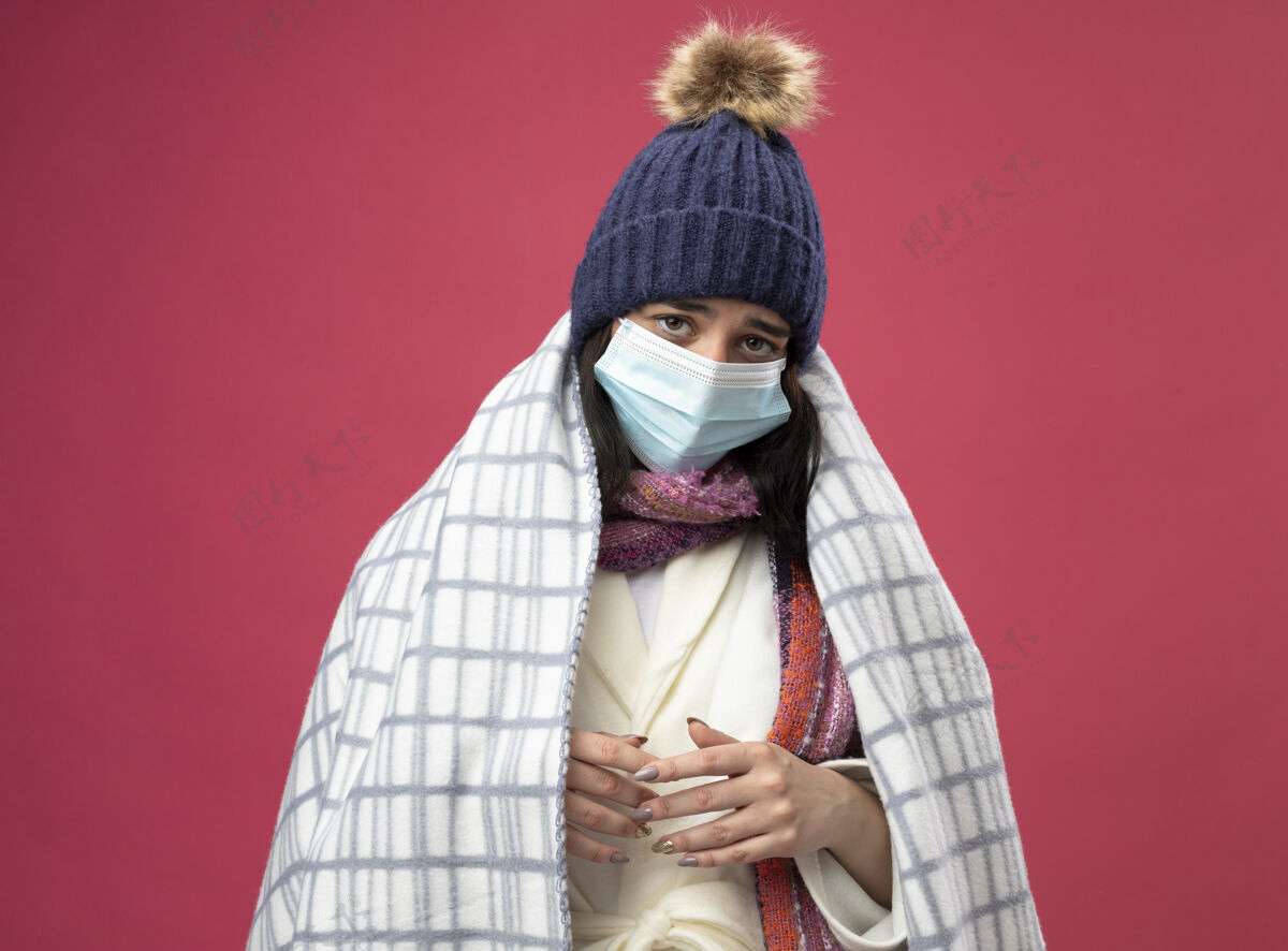 包裹穿着长袍 戴着冬日帽 戴着围巾 戴着方格布面具 双手合十 看着深红背景上的摄像机年轻疾病围巾