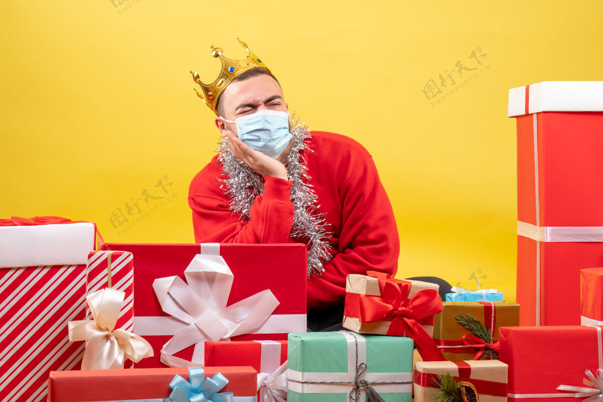 面具正面图：坐在圣诞节周围的年轻男性戴着黄色背景的面具出现人礼物漂亮