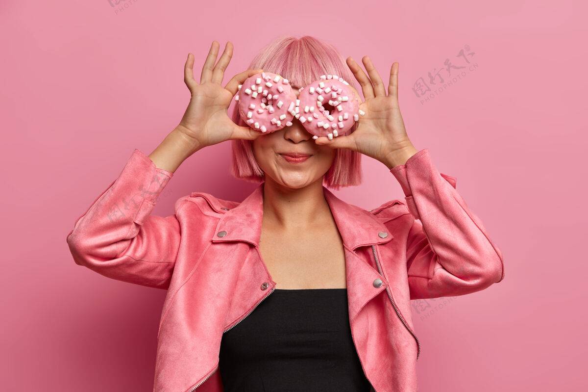 封面时尚粉发亚洲女孩的照片用美味的甜甜圈遮住眼睛 享受芳香可口的糖果 吃着釉面甜甜圈粉色假发食物乐趣