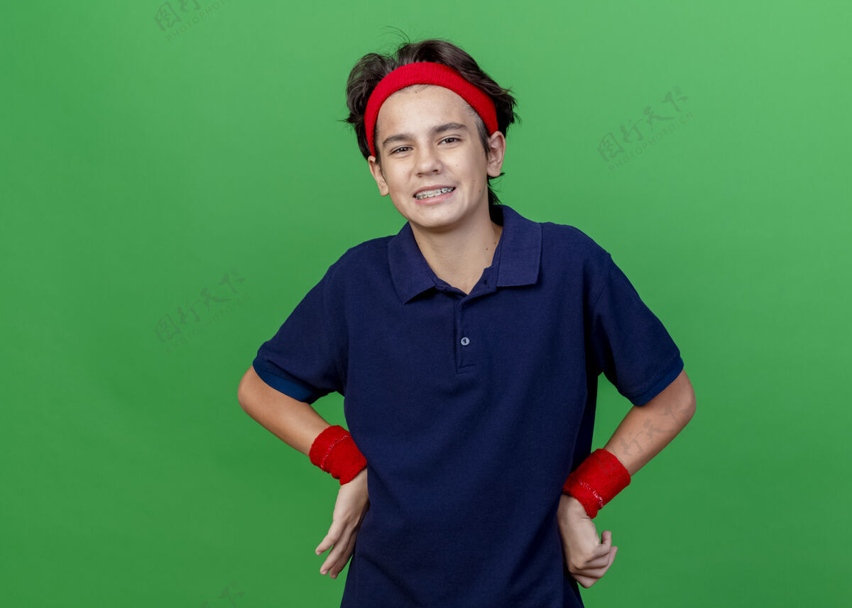 腰微笑的年轻帅气的运动男孩戴着头带和带牙套的腕带 手放在腰上 看着绿色背景上的相机 还有复制空间头带手微笑