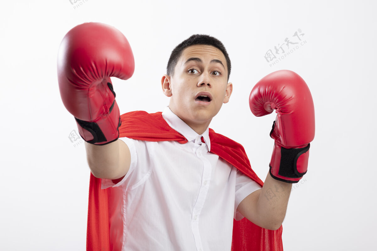 红令人印象深刻的年轻超级英雄男孩穿着红色斗篷盒手套看着一边保持手在空气中伸展另一只手对着相机隔离在白色背景上伸展白手套