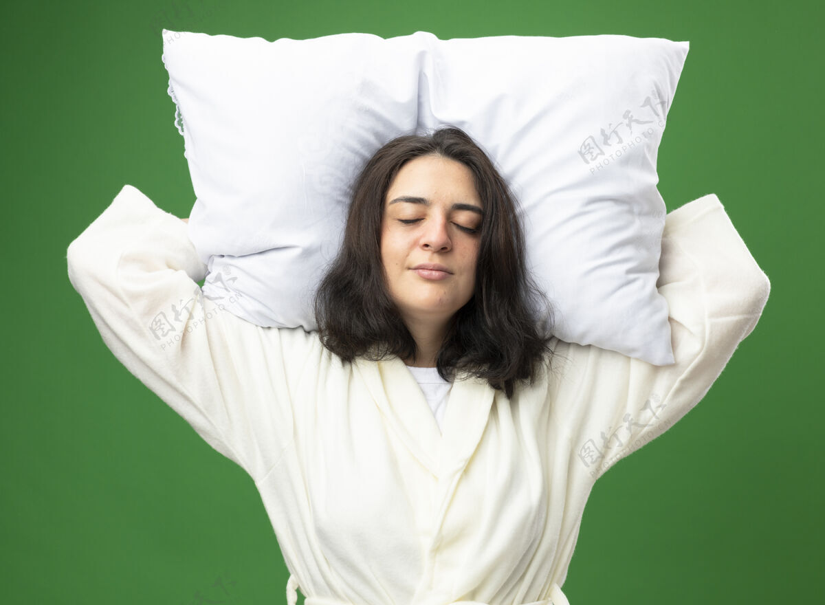穿年轻的白人病女孩穿着长袍抱着枕头在头下假装睡在绿色背景隔离假装抱着头