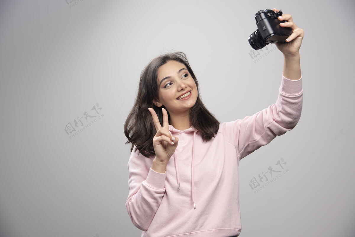 女人漂亮的年轻女子在灰色的墙上用相机自拍站着年轻人