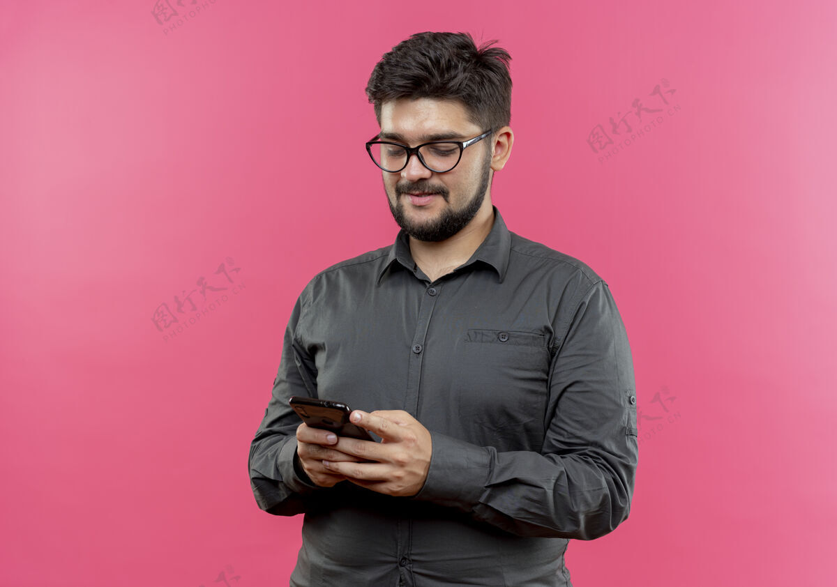 商人高兴的年轻商人戴着眼镜拿着手机看着隔离在粉红色墙上年轻人墙请戴上