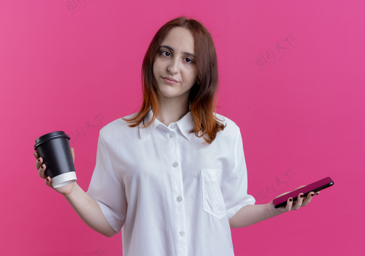 咖啡年轻的红发女孩拿着电话和咖啡孤立在粉红色的墙上年轻拿着粉色