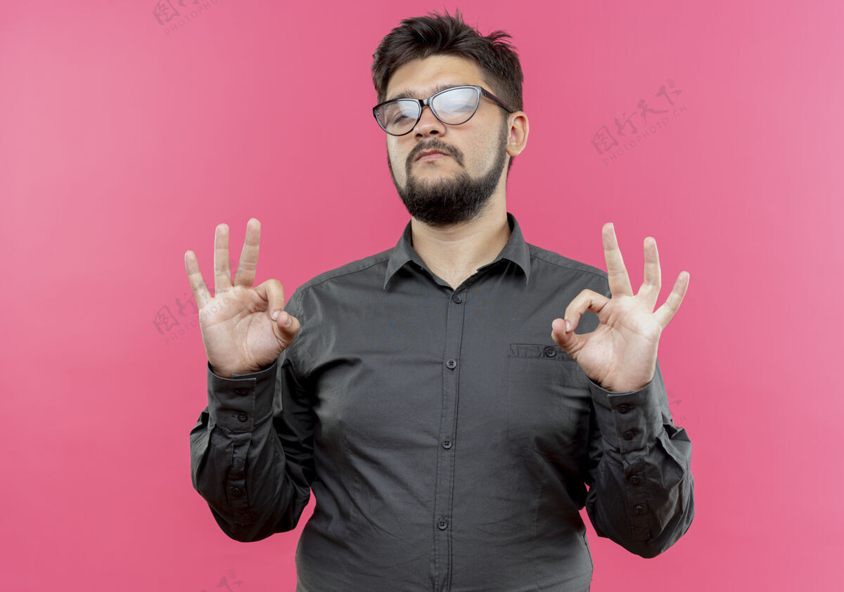 穿自信的年轻商人戴着眼镜 在粉红色的墙上显示出一种很好的姿态手势商人好的